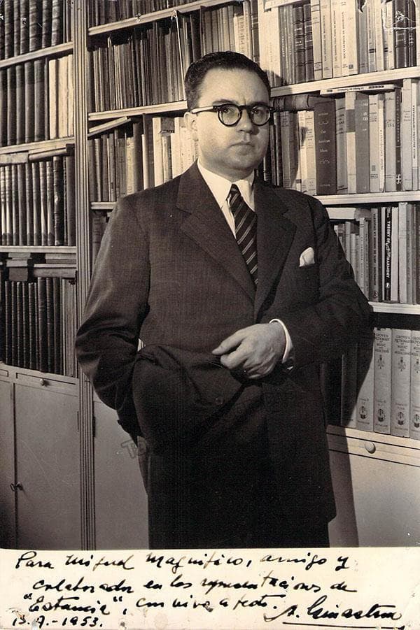 Alberto Ginastera standing in front of bookshelves of music scores
